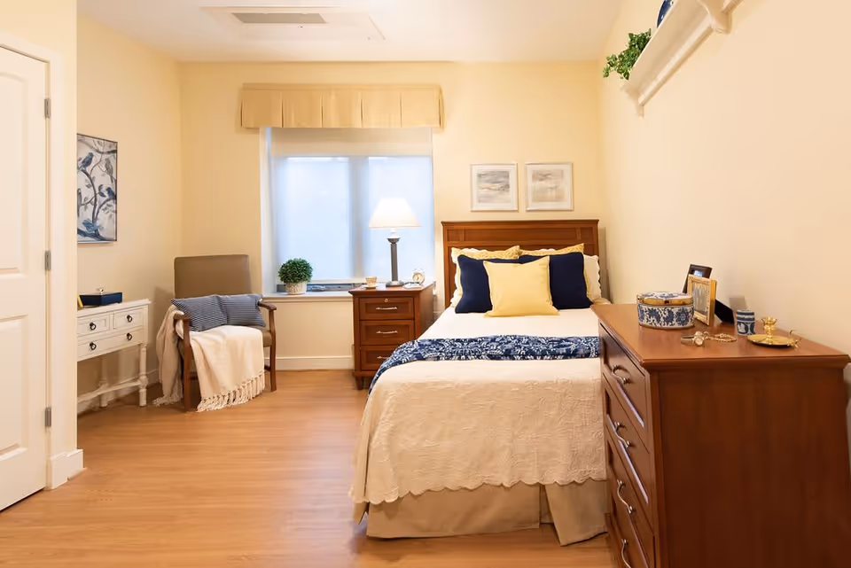 A cozy bedroom with a wooden bed featuring white and blue bedding and yellow and navy pillows. There is a wooden nightstand with a lamp and decorative items next to the bed, a wooden dresser with various decorative objects on the right, and a cushioned armchair with a throw blanket and pillow near a window with a beige valance. The walls are light beige, and there is a small white desk with drawers and a framed artwork on the left wall.