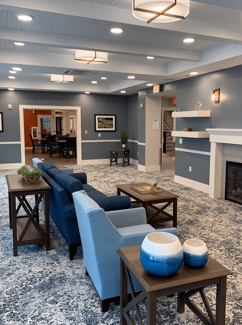 A spacious, well-lit lounge with blue upholstered sofas and armchairs, wooden side tables, patterned carpet, and a fireplace.