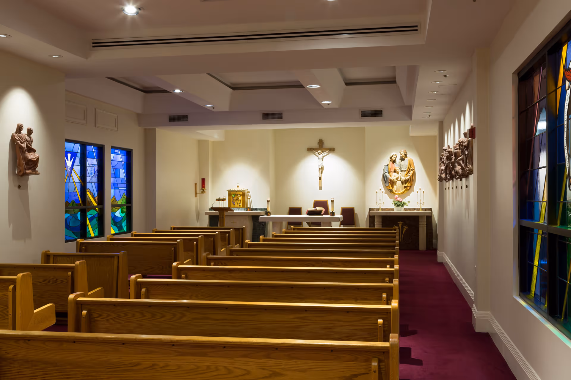 Interior view of a small chapel with wooden pews arranged in rows facing an altar. The altar area features religious statues, a crucifix on the wall, and candles. Stained glass windows with colorful designs are visible on the left and right walls. The floor is carpeted in dark red.