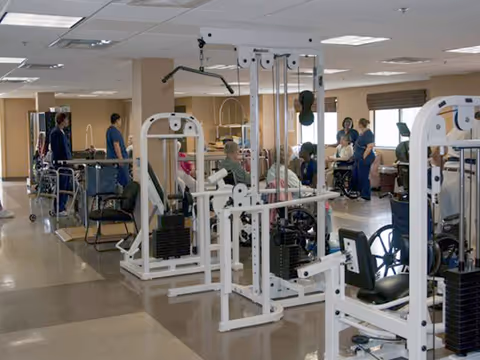 A rehabilitation center room with exercise equipment and several people, some seated in wheelchairs and others standing, engaging in physical therapy activities.
