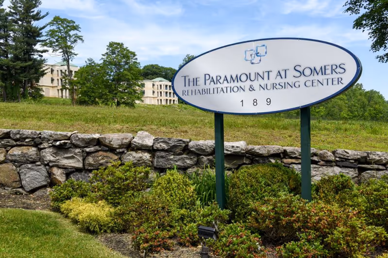 Oval sign for The Paramount at Somers Rehabilitation & Nursing Center near a stone wall.