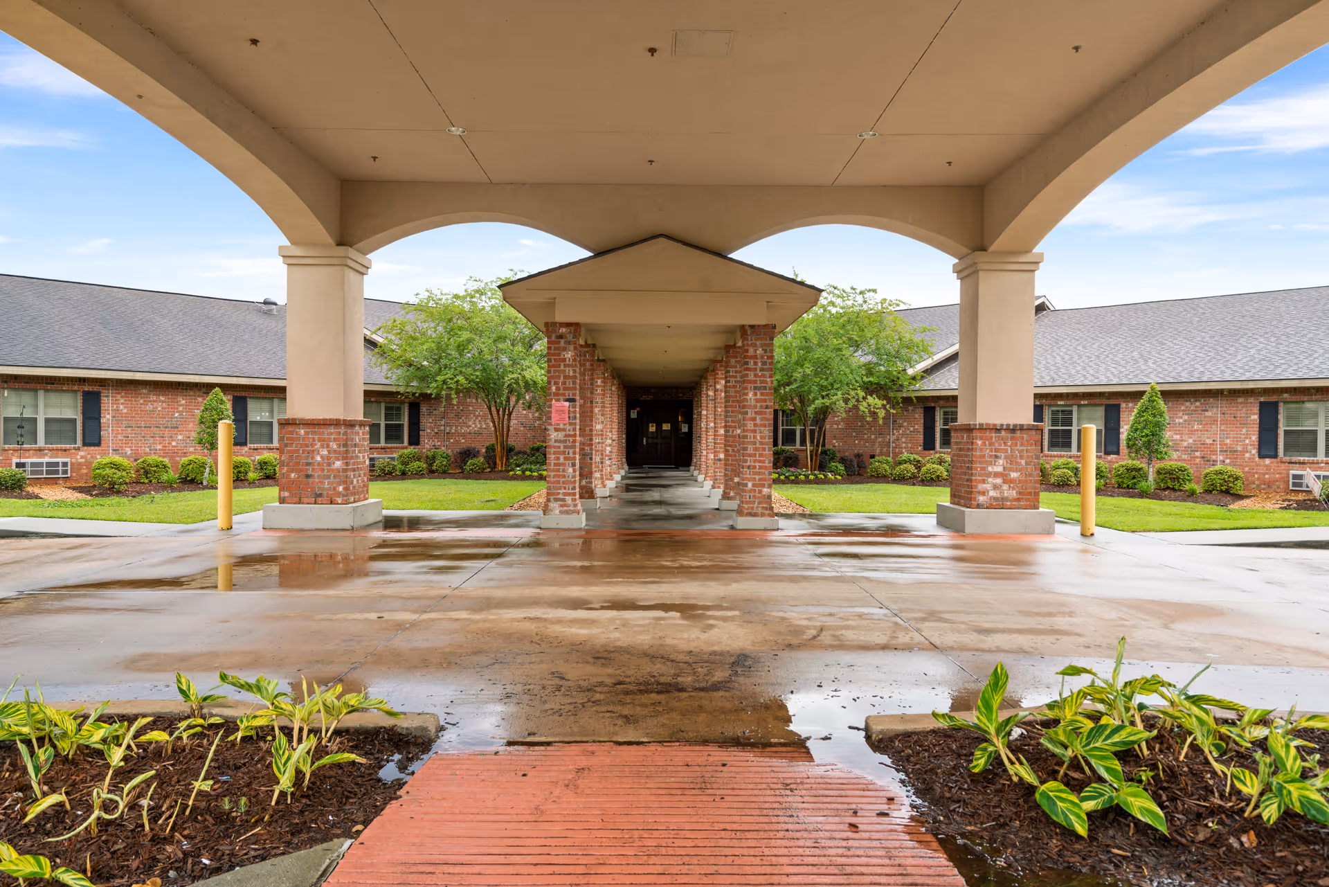 Covered entrance driveway to a brick building with a walkway supported by brick columns, surrounded by green shrubs and small trees under a partly cloudy sky.