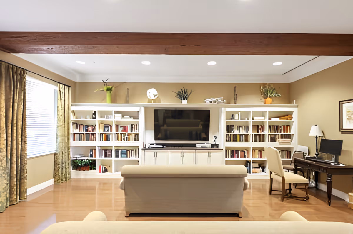 A spacious living room with a large white bookshelf filled with books and decorative items, a flat-screen TV in the center, a beige sofa facing the TV, a wooden desk with a computer and chair on the right side, and large windows with green curtains on the left side. The room has wooden flooring and a wooden beam across the ceiling.