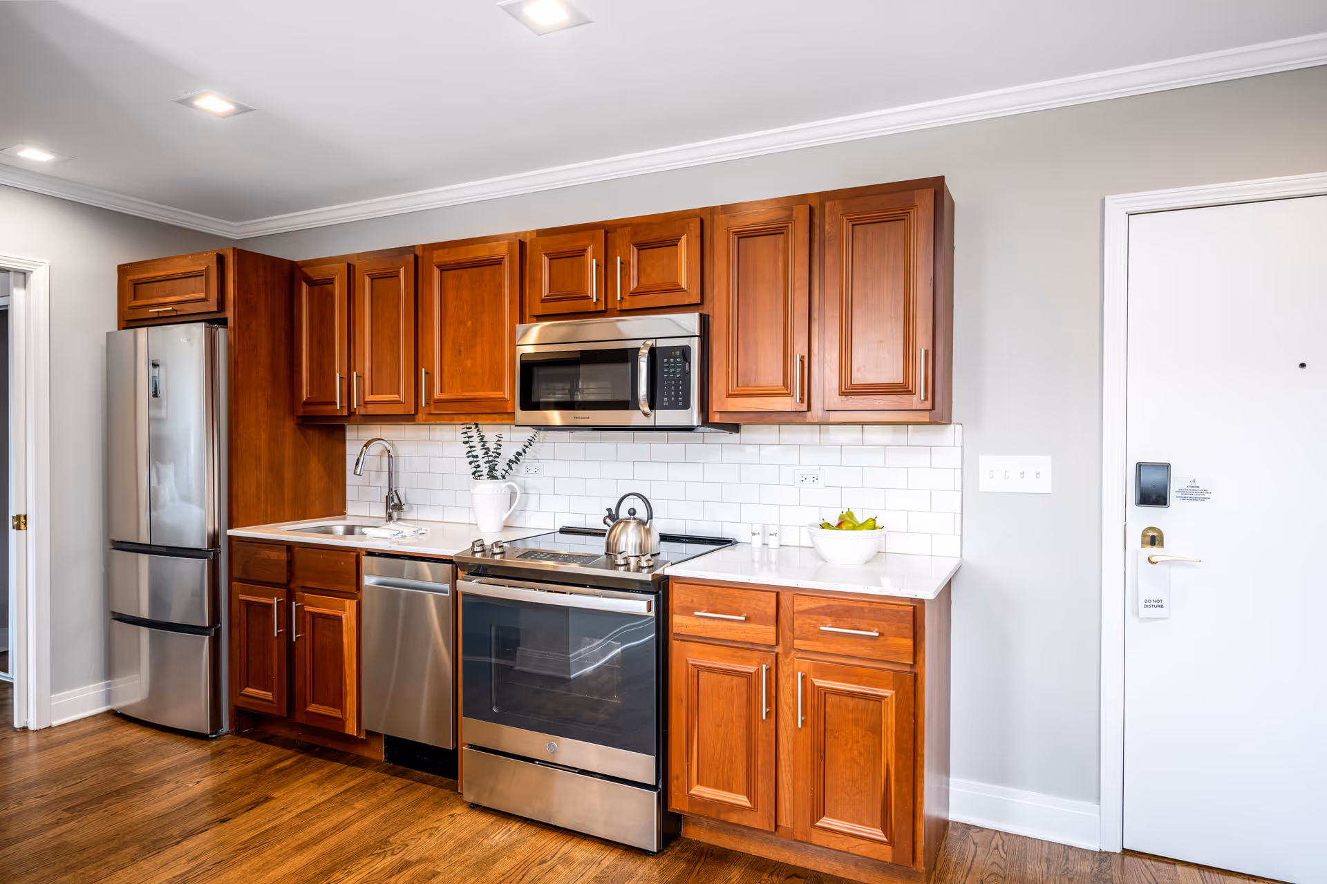 A modern kitchen with wooden cabinets, stainless steel refrigerator, dishwasher, oven, and microwave. The backsplash is white subway tile, and there is a white countertop with a bowl of fruit and a white vase with greenery. The floor is wooden, and there is a white door to the right.