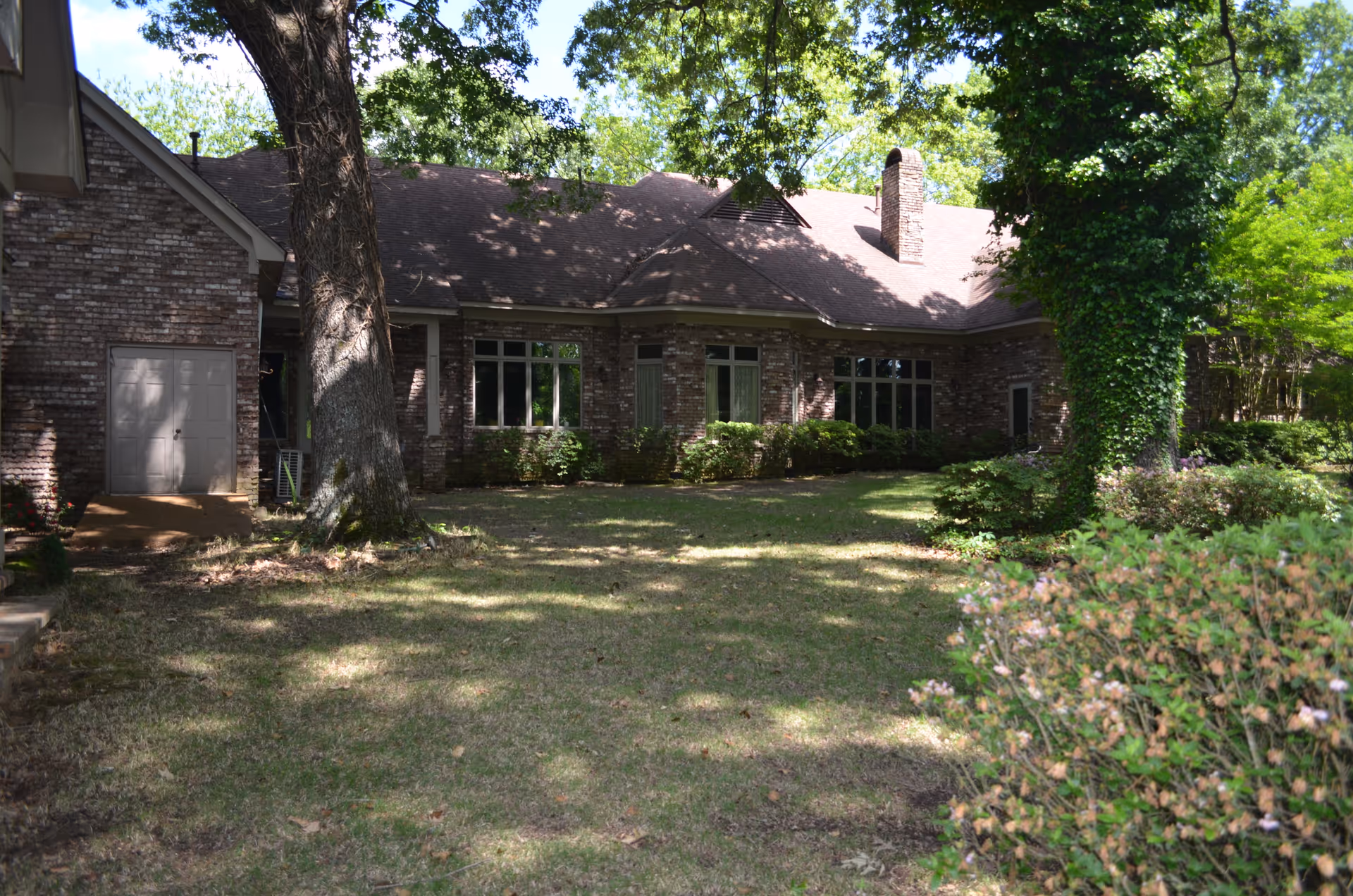 View of a brick building with multiple windows surrounded by trees and bushes, with a grassy lawn in the foreground under a sunny sky.