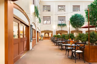 Atrium-style indoor dining area with tables and chairs, potted trees, string lights, and surrounding multi-story windows.
