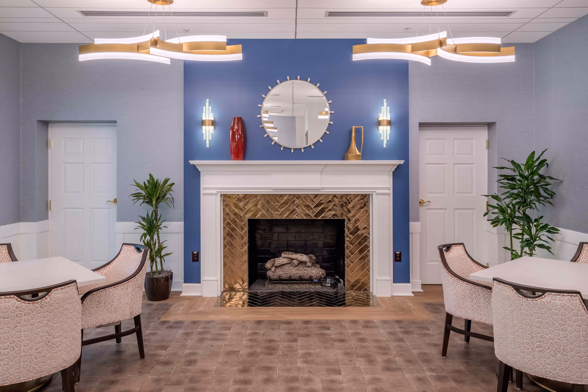 A cozy interior room with a central fireplace featuring a white mantel and a gold-tiled surround. Above the fireplace is a round mirror flanked by two modern wall sconces. On the mantel are decorative vases. The walls are painted blue behind the fireplace and light gray on the sides. There are two white doors on either side of the fireplace, with potted plants nearby. Two tables with patterned upholstered chairs are positioned on either side of the room. Modern ceiling lights hang above.