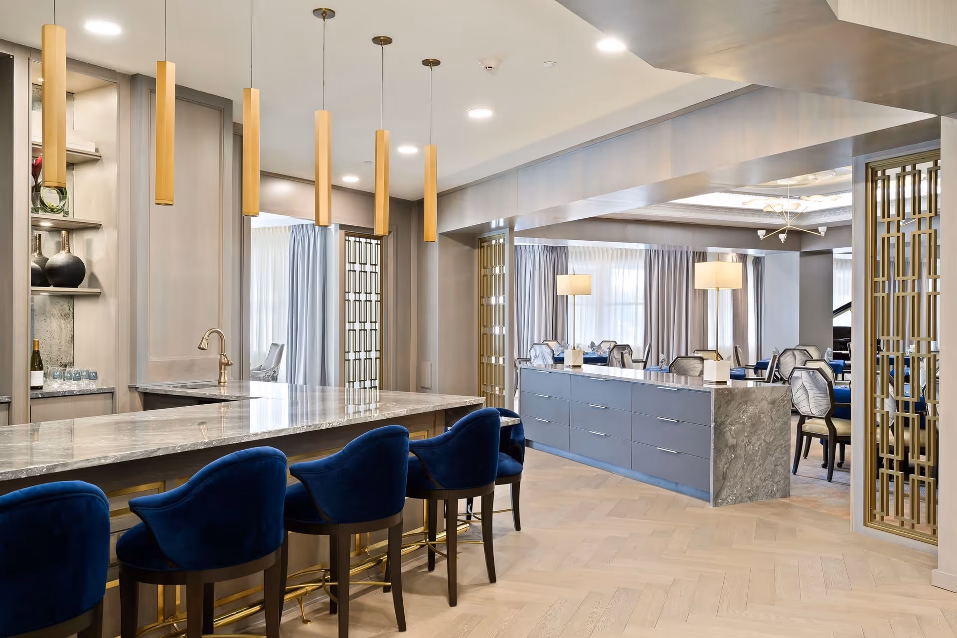 A modern and elegant interior space featuring a marble countertop bar with five blue velvet bar stools. Above the bar hang five cylindrical pendant lights with a gold finish. In the background, there is a large room with multiple tables and chairs, soft lighting from table lamps, and large windows with light-colored curtains. The space has light wood flooring and decorative gold metal room dividers.