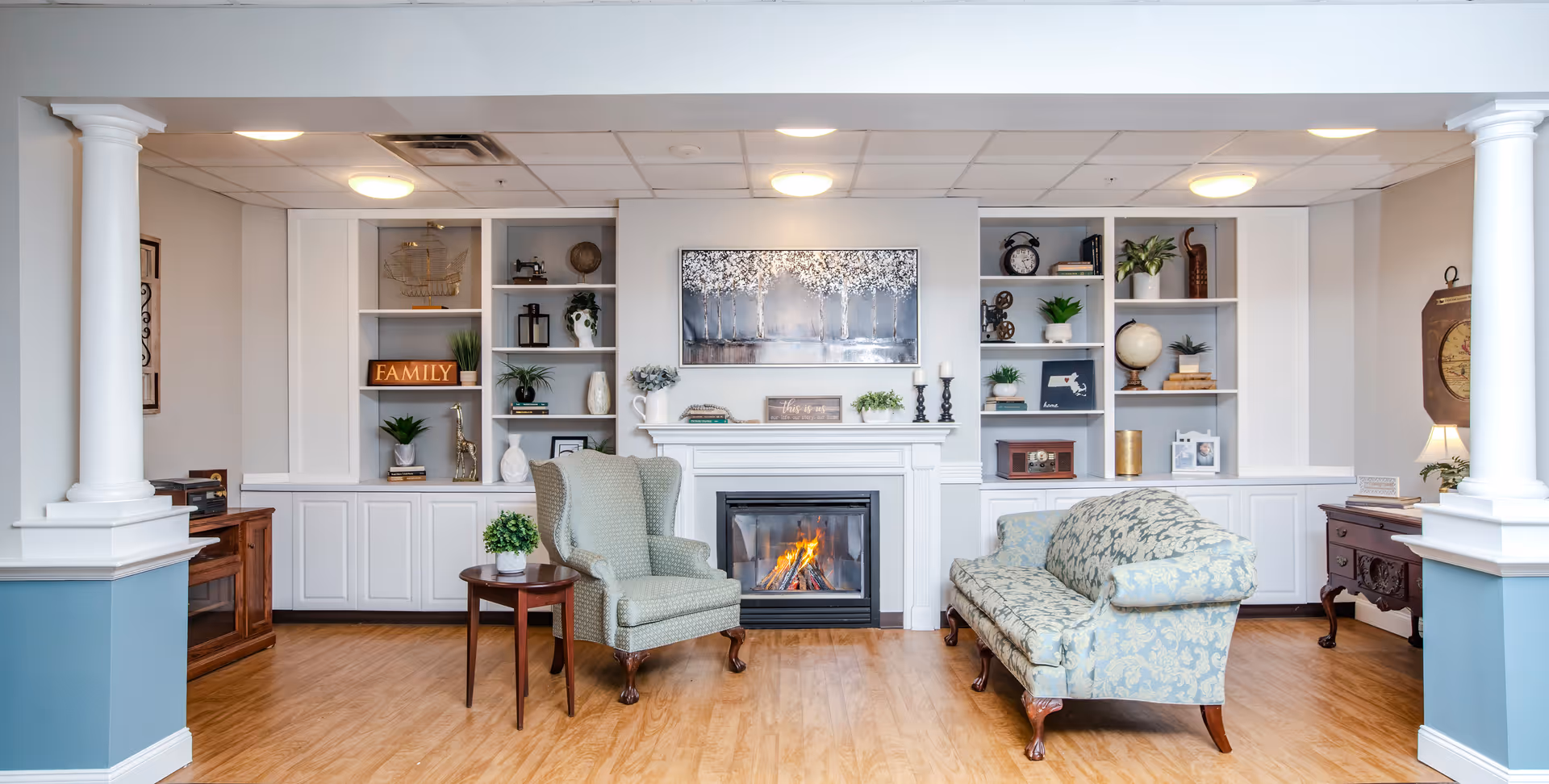 A cozy senior living room with a lit fireplace centered between built-in white shelves filled with decorative items and plants. There is a patterned armchair with a small round wooden side table holding a potted plant on the left, and a floral upholstered sofa on the right. The room features wooden flooring, white columns, and soft overhead lighting.