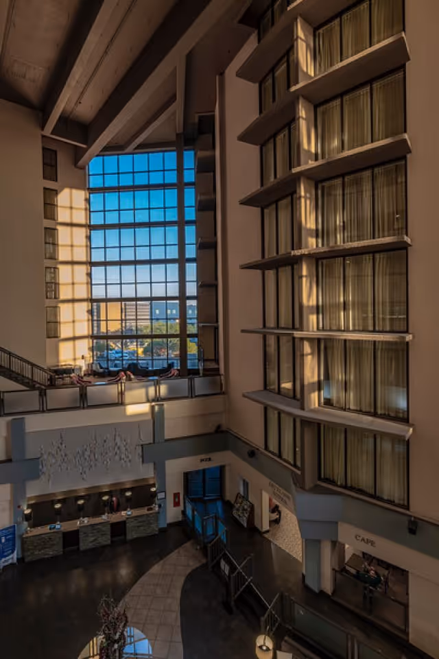 Interior view of a multi-story senior living facility lobby with large windows letting in natural light. The lobby features a reception desk, seating areas, and signs for a cafe and other amenities. The architecture includes tall ceilings and multiple floors with windows overlooking the lobby.
