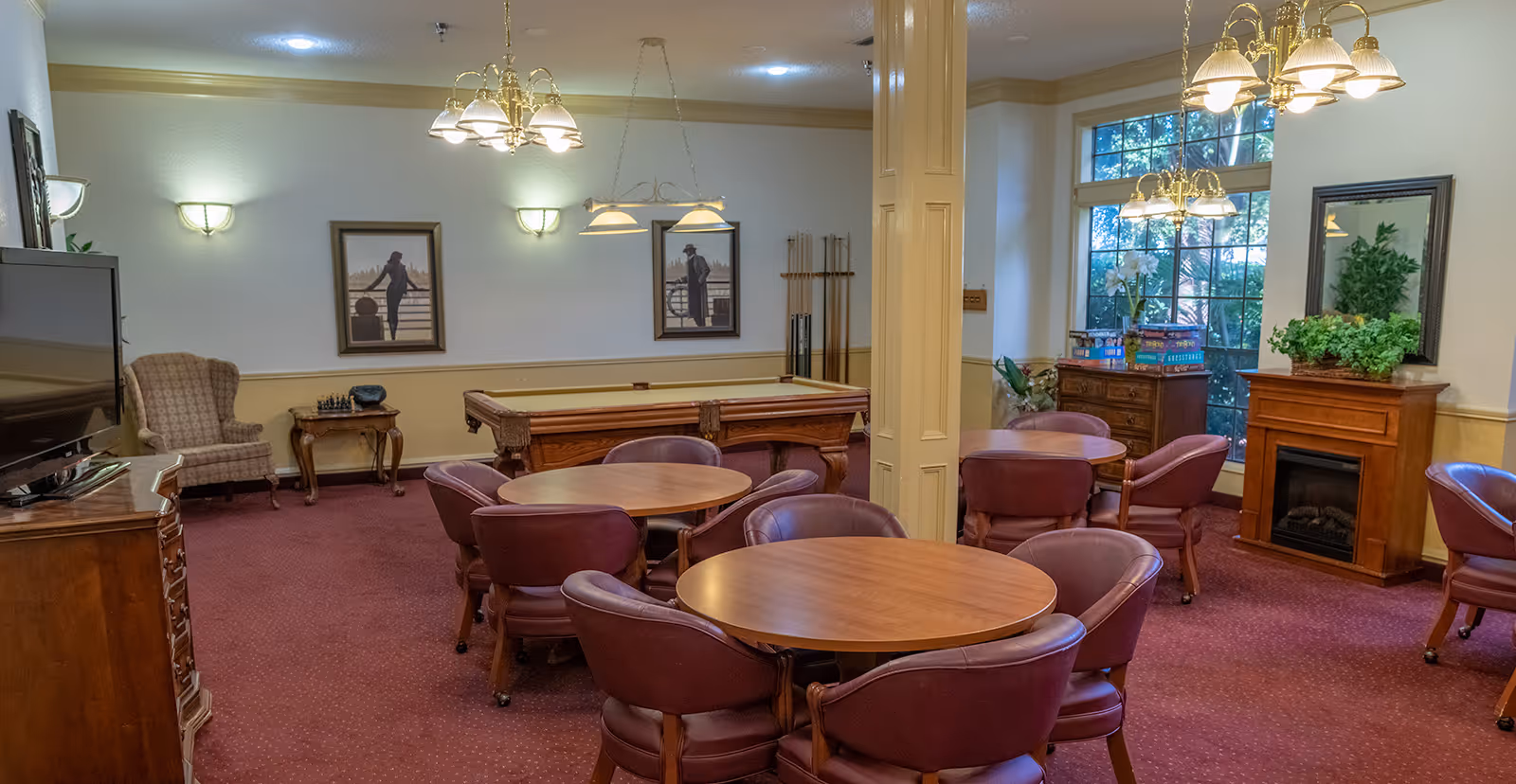 A cozy common room with round wooden tables surrounded by maroon leather chairs, a pool table in the background, a large window letting in natural light, a fireplace with a plant on the mantel, framed pictures on the walls, and warm overhead lighting fixtures.