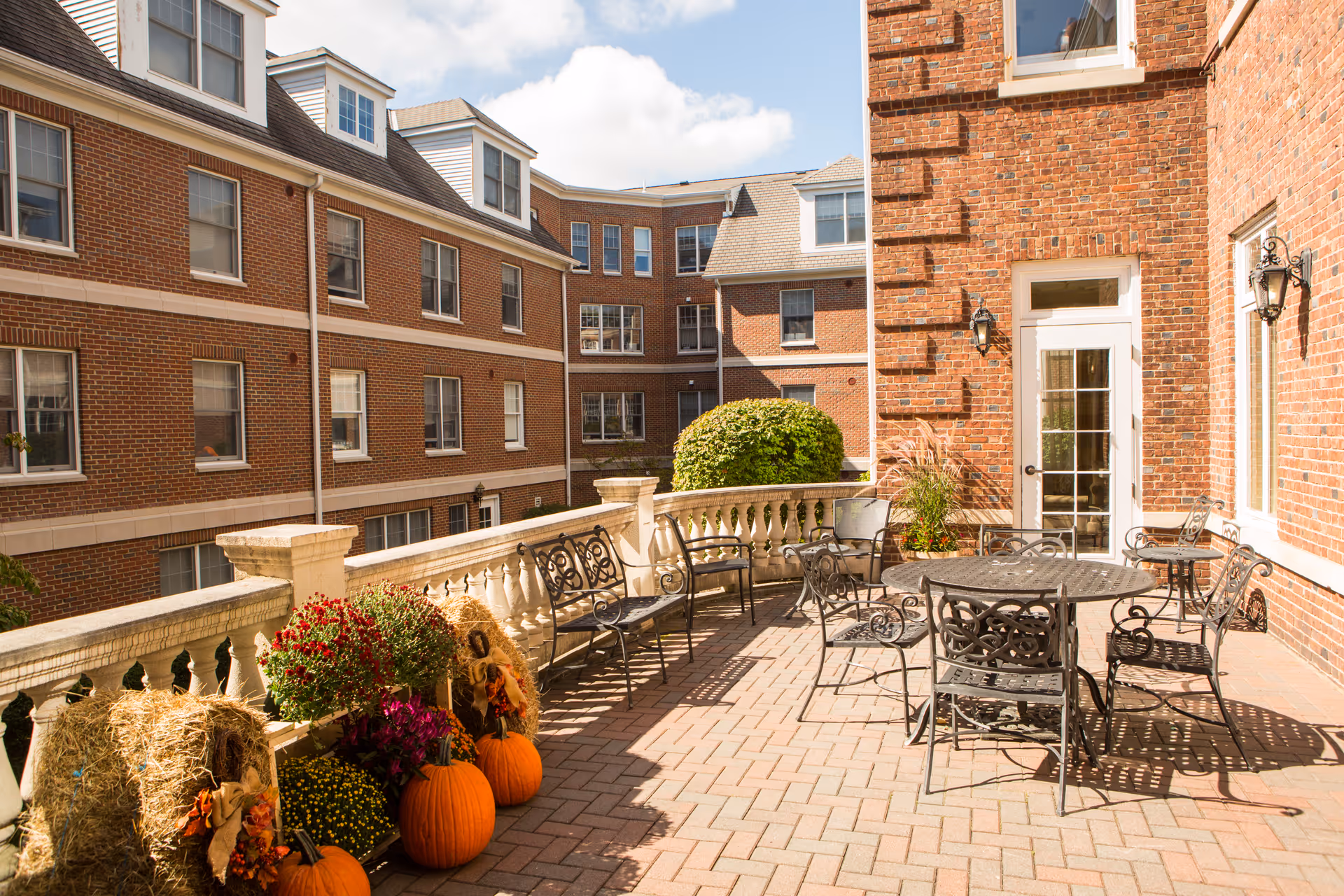 Sunny brick terrace with wrought-iron tables and chairs, seasonal pumpkins and potted plants beside a multi-story brick building.