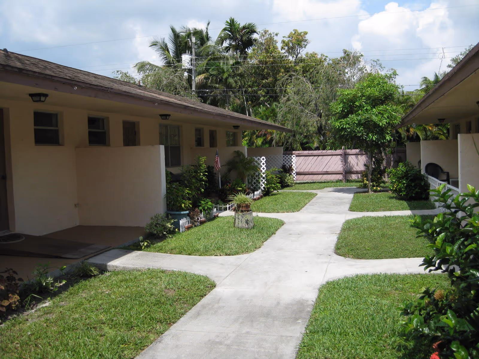 Outdoor courtyard area of a senior living facility with a concrete walkway intersecting through green grass and small garden beds. The courtyard is surrounded by single-story buildings with beige walls and brown roofs. There are various plants, shrubs, and small trees along the walkway and near the buildings. The sky is partly cloudy.