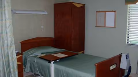 A small hospital-style bedroom with a single bed, wardrobe, and window in a senior facility.