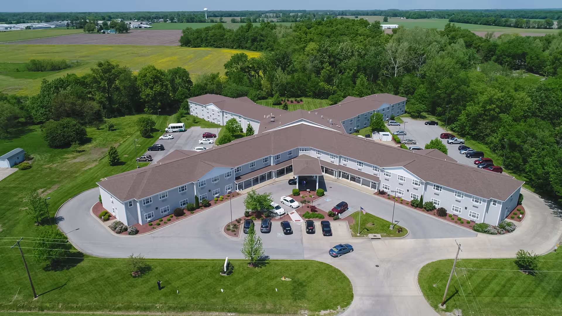 Aerial view of Heritage Woods of Benton, a large senior living facility surrounded by green trees and fields. The building has a brown roof and light-colored exterior walls, with multiple parking areas and cars parked around it. The facility is set in a rural area with open land and trees in the background.