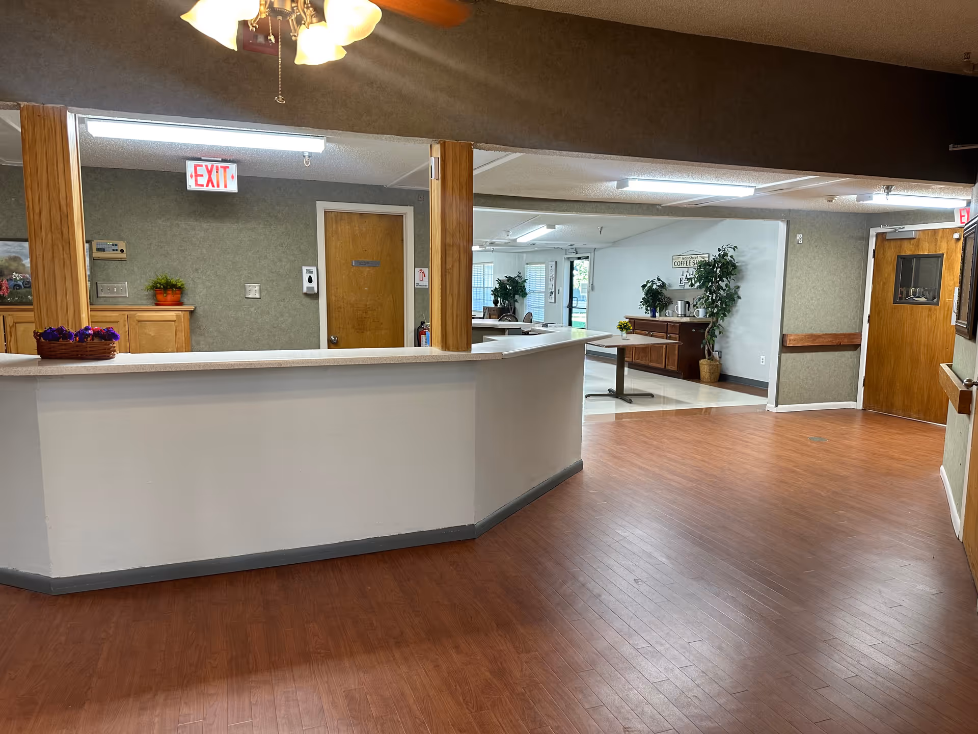 Interior view of a senior living facility reception area with a white counter, wooden floor, and wooden doors. There are plants and a table in the background, with exit signs above two doors.