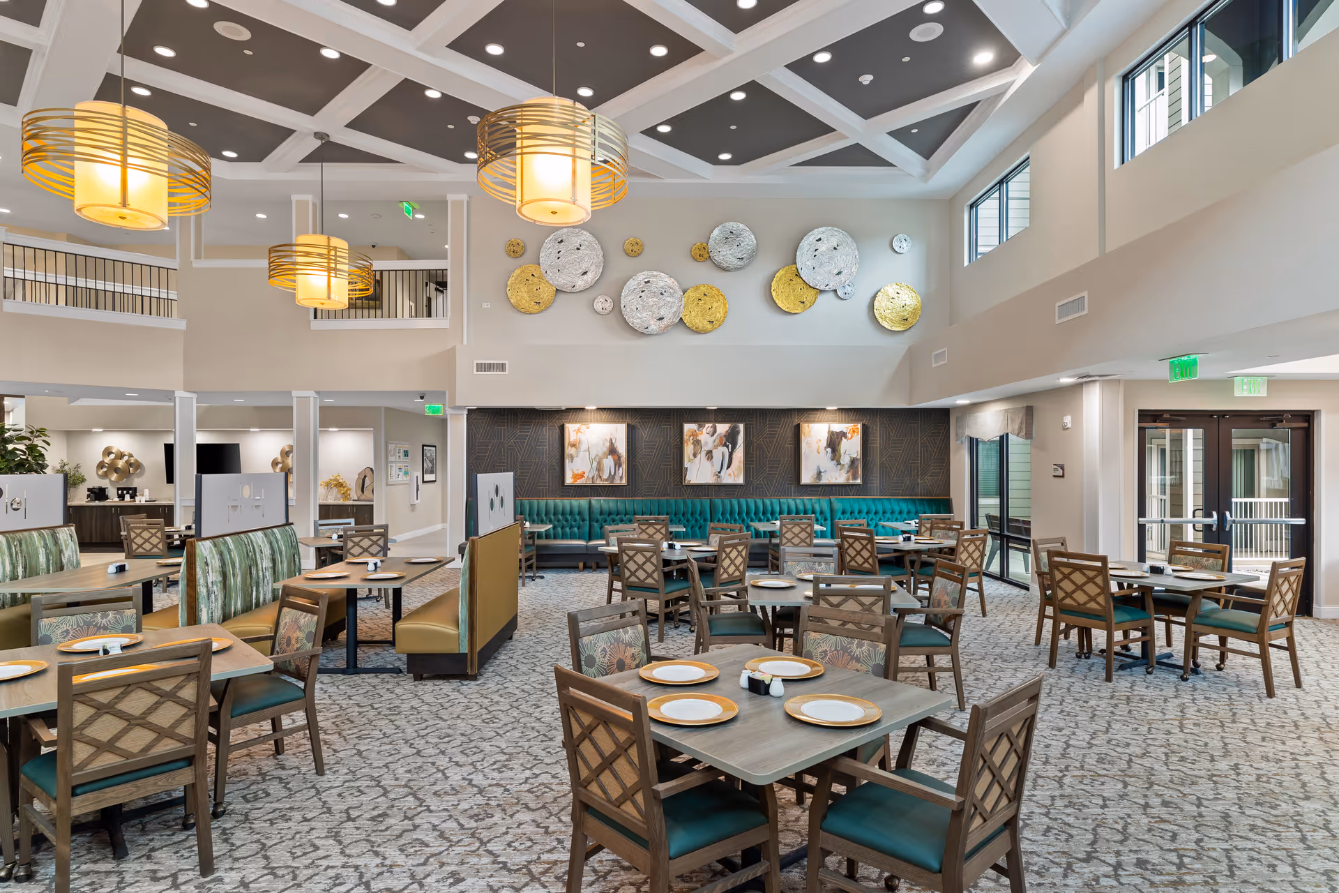 Spacious dining room in a senior living facility with multiple wooden tables and chairs arranged neatly. The room features high ceilings with modern pendant lights, large windows allowing natural light, decorative wall art, and a teal upholstered bench along one wall. The carpet has a patterned design, and the overall ambiance is bright and welcoming.