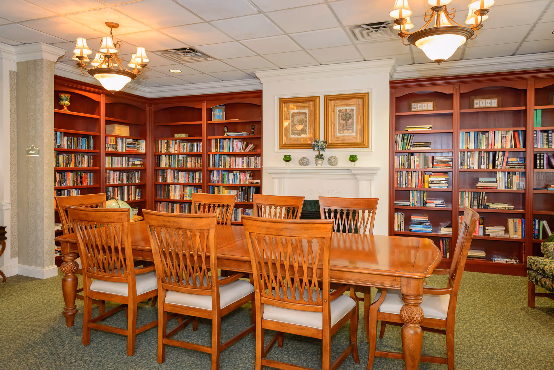 A cozy library room with wooden bookshelves filled with books, a large wooden table surrounded by eight matching chairs with cushioned seats, two framed pictures above a white fireplace mantel, and two ceiling light fixtures providing warm lighting.