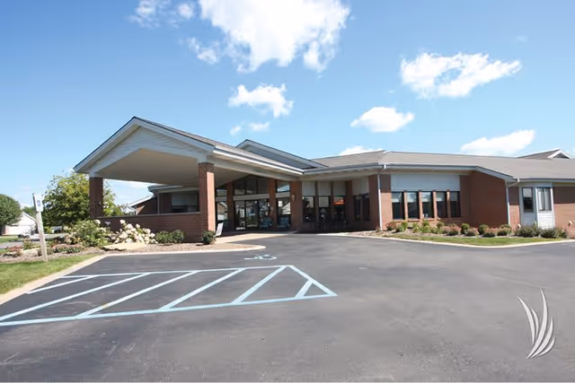 Exterior view of a single-story brick building with a covered entrance and large windows under a blue sky with scattered clouds. The parking lot in front has marked handicap parking spaces and landscaped areas with shrubs and flowers.