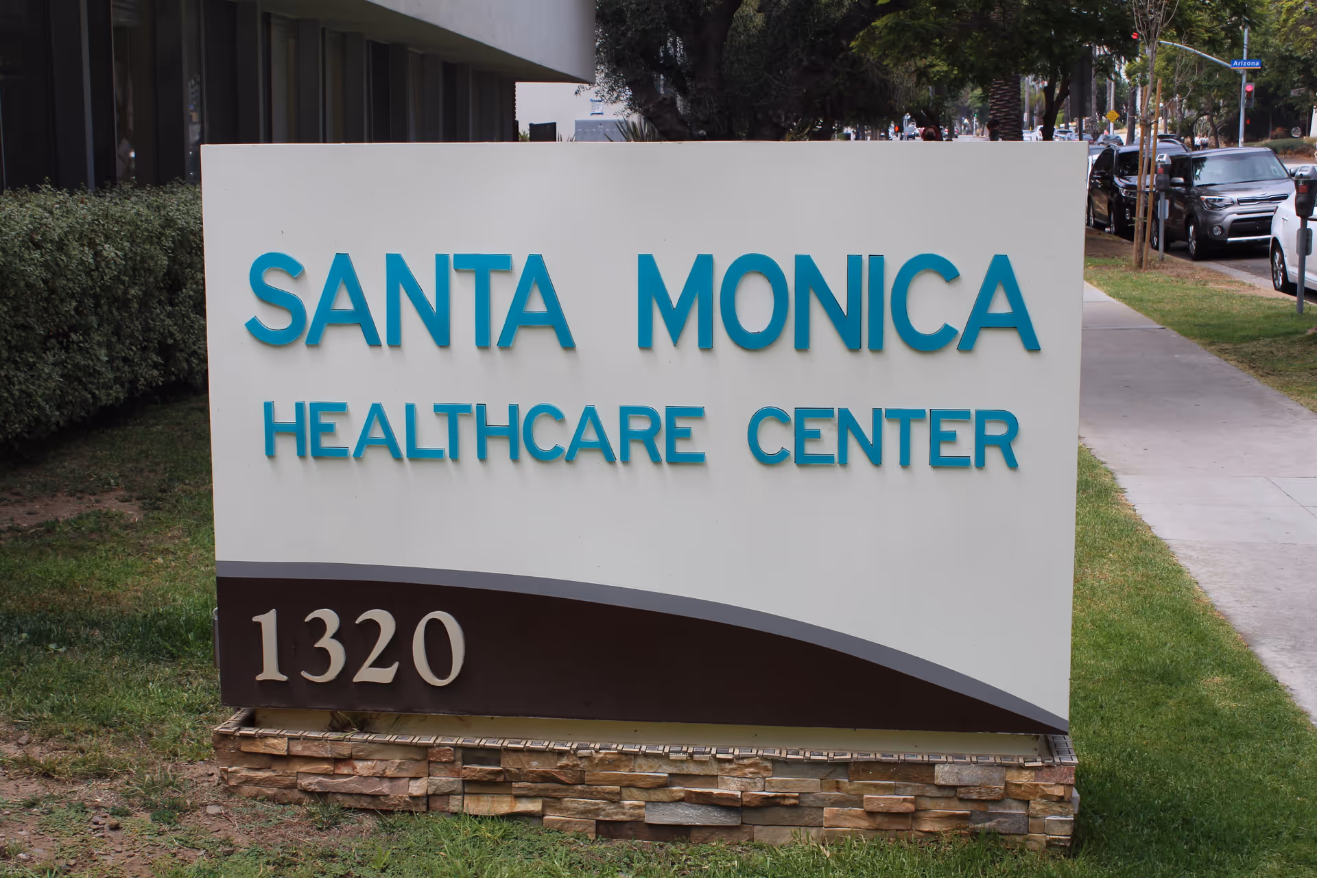 Outdoor sign for Santa Monica Healthcare Center with the address number 1320, situated on a grassy area next to a sidewalk and street with parked cars.