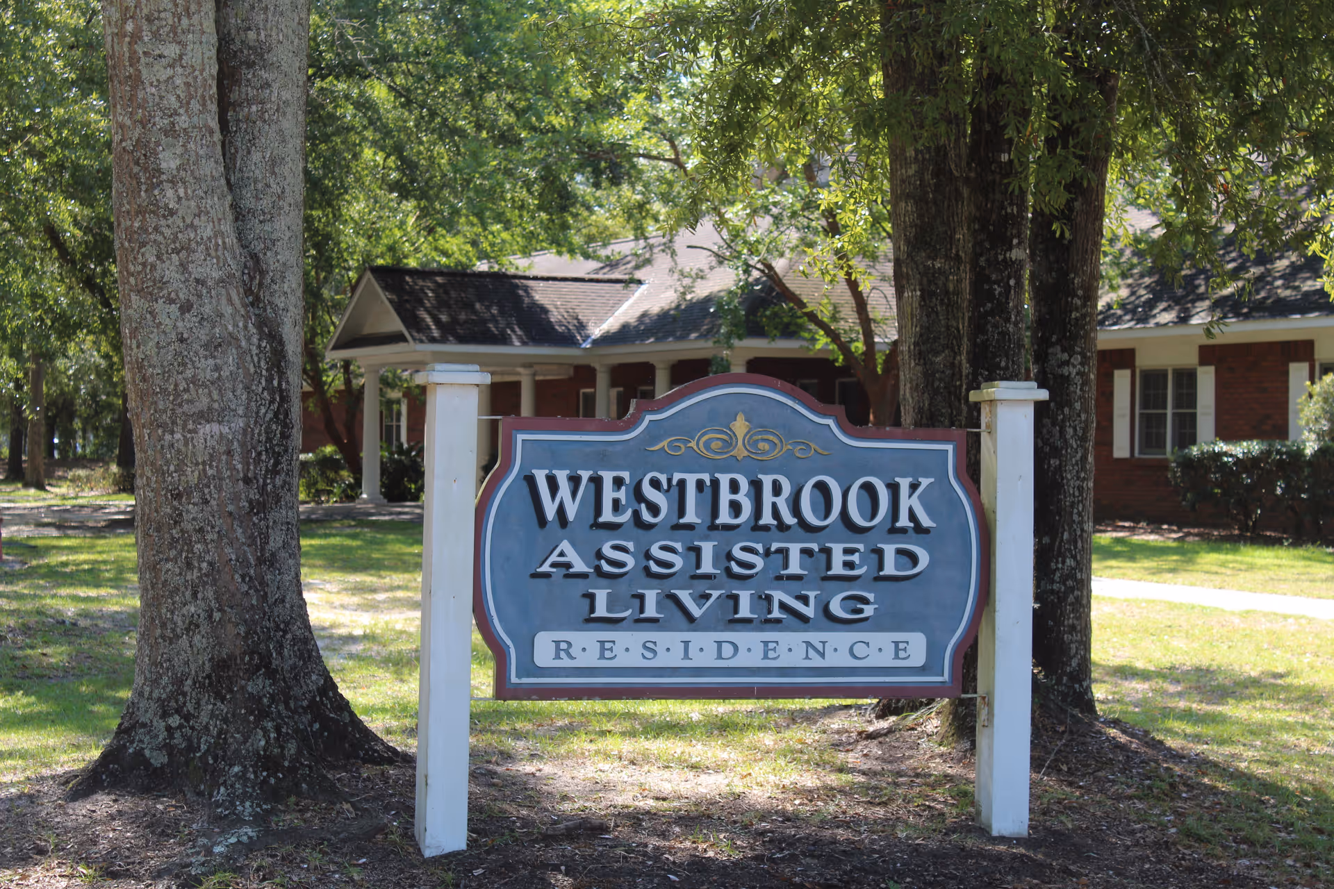 A sign for Westbrook Assisted Living Residence is displayed outdoors between two large trees, with a brick building and green lawn visible in the background.