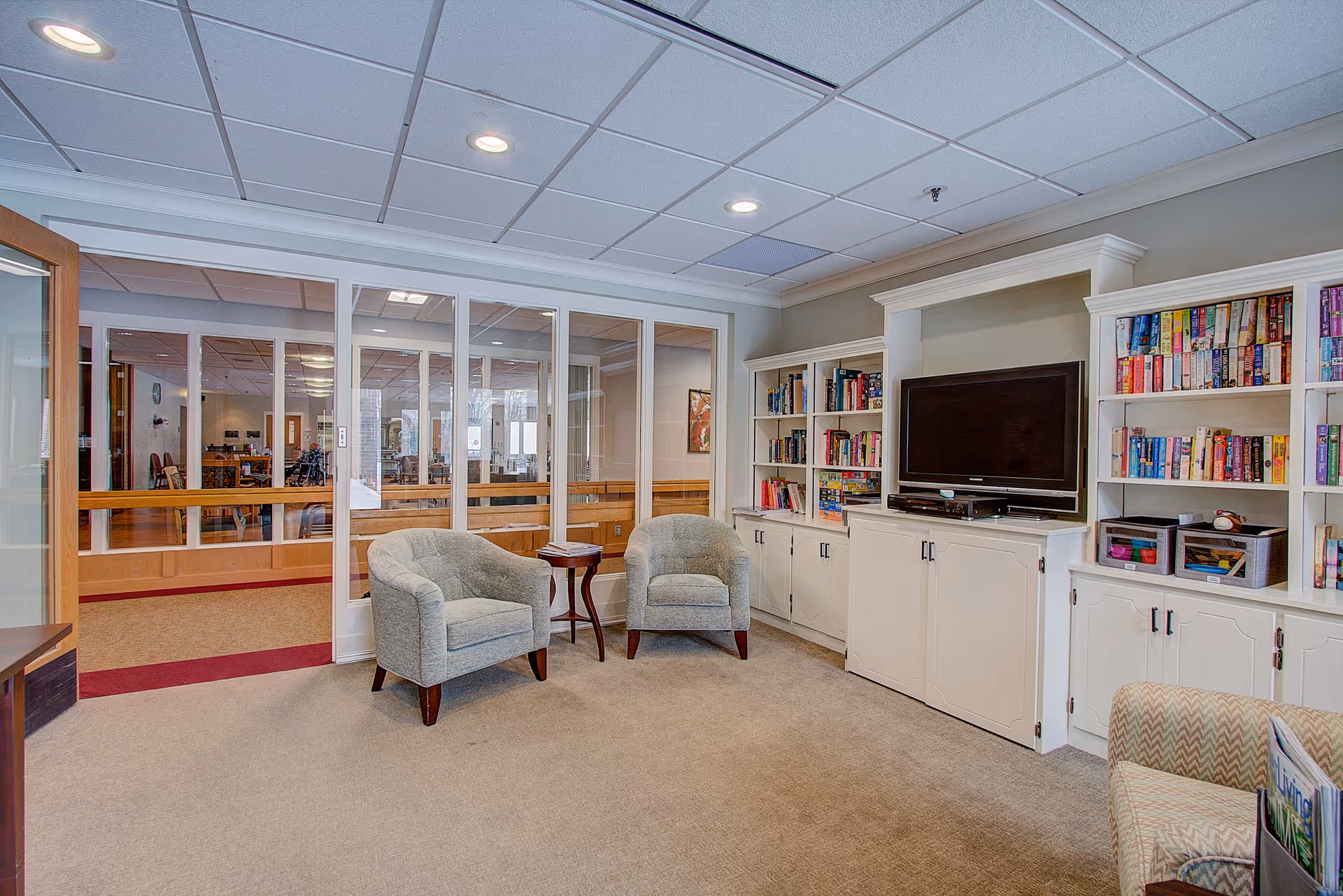 A cozy senior living community lounge area with two gray armchairs and a small wooden side table between them. The room features built-in white bookshelves filled with books and a flat-screen TV mounted on a cabinet. The space has a beige carpet and a drop ceiling with recessed lighting. Glass sliding doors separate this room from another area with more seating and tables.