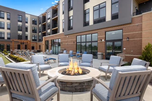 A modern outdoor courtyard with a central stone fire pit surrounded by cushioned lounge chairs in front of a multi-story building.