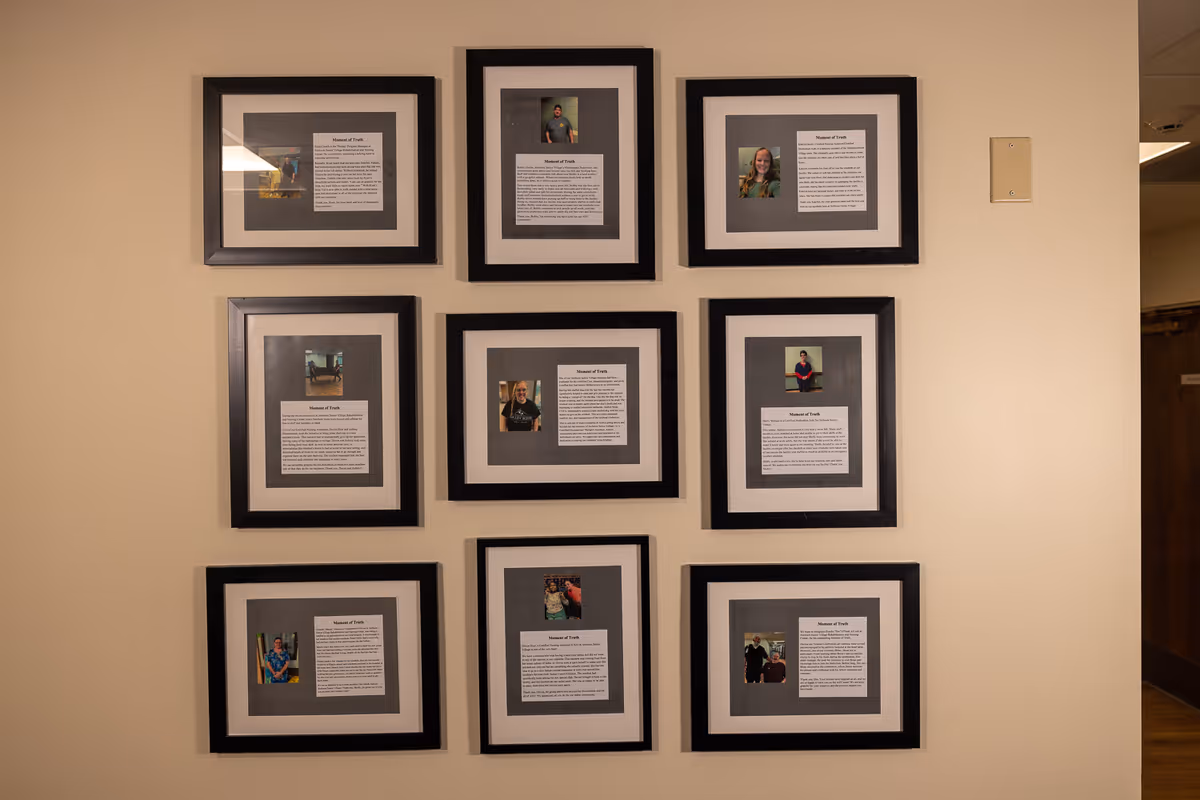 A beige wall with nine black-framed photos and text plaques arranged in a grid. Each frame contains a small portrait photo above a block of text, likely describing the person or their story.