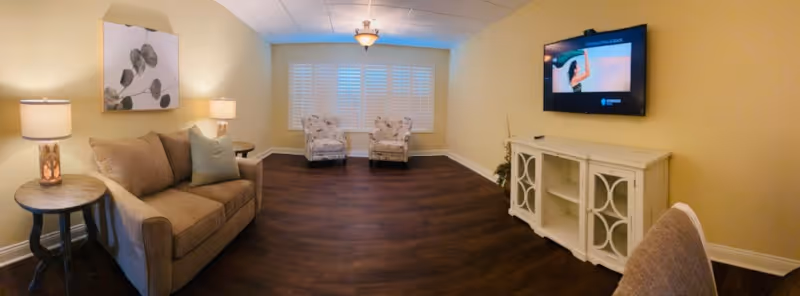 A cozy living room with a beige sofa, two patterned armchairs near a window with white shutters, a white TV stand with a flat-screen TV mounted above it, and warm lighting from table lamps and a ceiling light fixture. The room has dark wood flooring and light yellow walls.