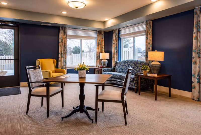 A cozy senior living common area with a wooden table and two chairs in the center, a yellow armchair to the left, and a blue patterned sofa to the right. There are two side tables with lamps and a flower vase on the table. Large windows with floral curtains let in natural light, and the walls are painted dark blue.