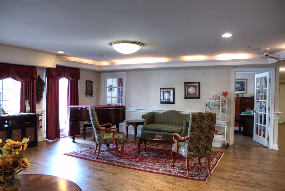 A cozy assisted living facility common area with wooden flooring, a green upholstered sofa, two floral-patterned armchairs, a wooden coffee table on a red patterned rug, a grand piano near windows with red curtains, framed artwork on the walls, and a birdcage near an open door leading to an office.