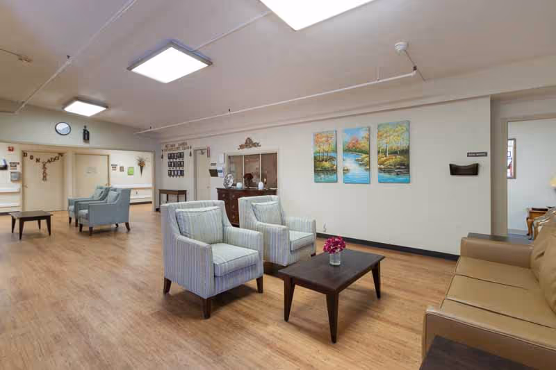A spacious and well-lit common area in a nursing and rehabilitation center featuring wooden flooring, two striped armchairs and a dark wooden coffee table with a small flower vase, a tan leather sofa, and colorful landscape paintings on the wall. Additional seating and decor are visible in the background.