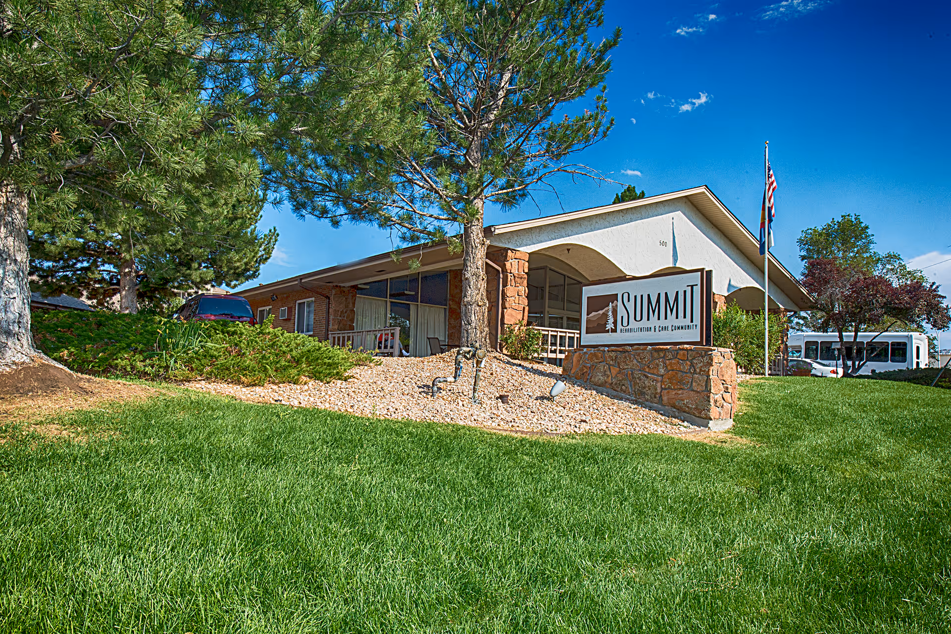 Front exterior of a low one-story care facility with a 'SUMMIT Rehabilitation & Care Community' sign, flagpoles, trees, and a manicured lawn.