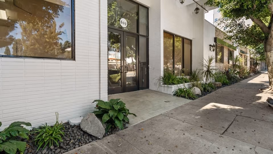 Street-level view of a white brick building entrance with glass double doors numbered 619, large windows, landscaping, and a sidewalk.