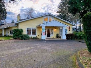 Exterior view of a single-story yellow building with white trim and a covered entrance. The building is surrounded by trees and bushes, with a paved driveway leading up to the entrance. The sky is partly cloudy.