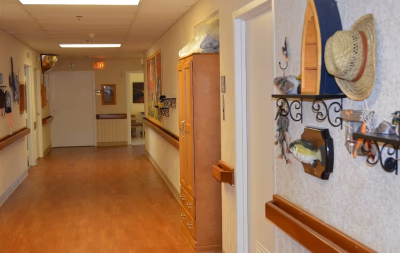 A decorated nursing home hallway with wood-look flooring, handrails, cabinets, and wall shelves holding hats and nautical decor.