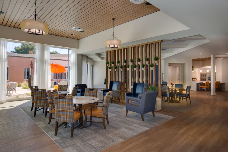 A spacious and well-lit common area in a senior living facility featuring multiple seating arrangements with plaid and solid upholstered chairs around round tables. Large windows with sheer white curtains allow natural light to fill the room, and an orange patio umbrella is visible outside. The ceiling has wooden paneling with modern light fixtures, and a wooden slatted divider with small potted plants separates the seating areas. In the background, a kitchen area with bar stools and pendant lights is visible.