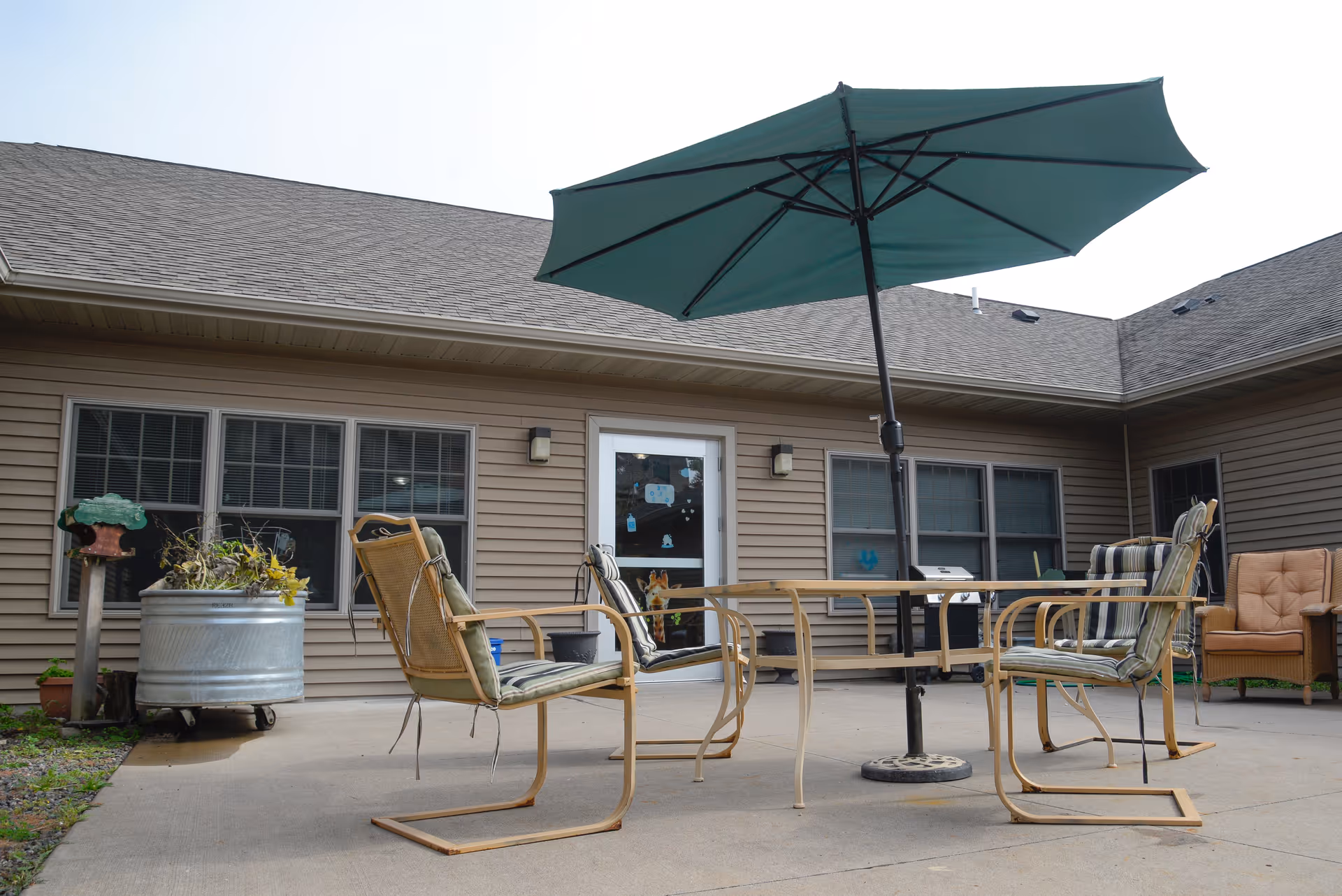 Outdoor patio area at VitaCare Living of Proctor with a round glass table, four cushioned chairs, and a large green umbrella providing shade. The patio is adjacent to a beige building with multiple windows and a door. There is a large metal planter with plants and a cushioned armchair in the corner.