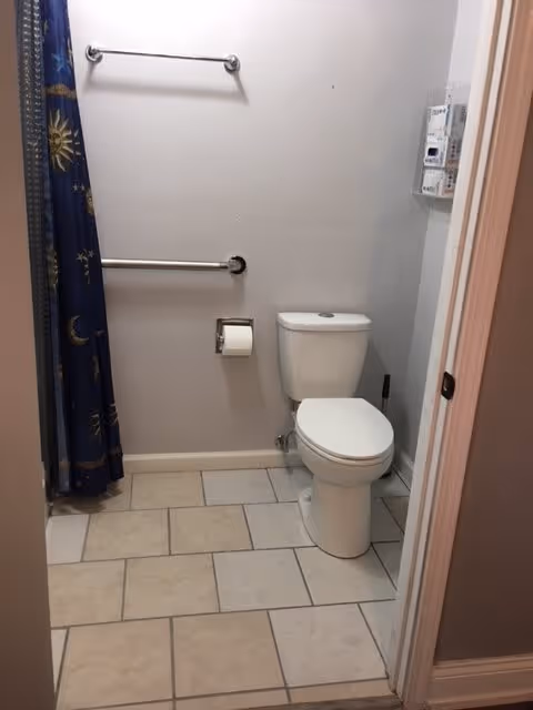 A small bathroom with a white toilet, a toilet paper holder mounted on the wall, two metal grab bars, and a blue shower curtain with sun and moon patterns. The floor is tiled with beige and light brown tiles.