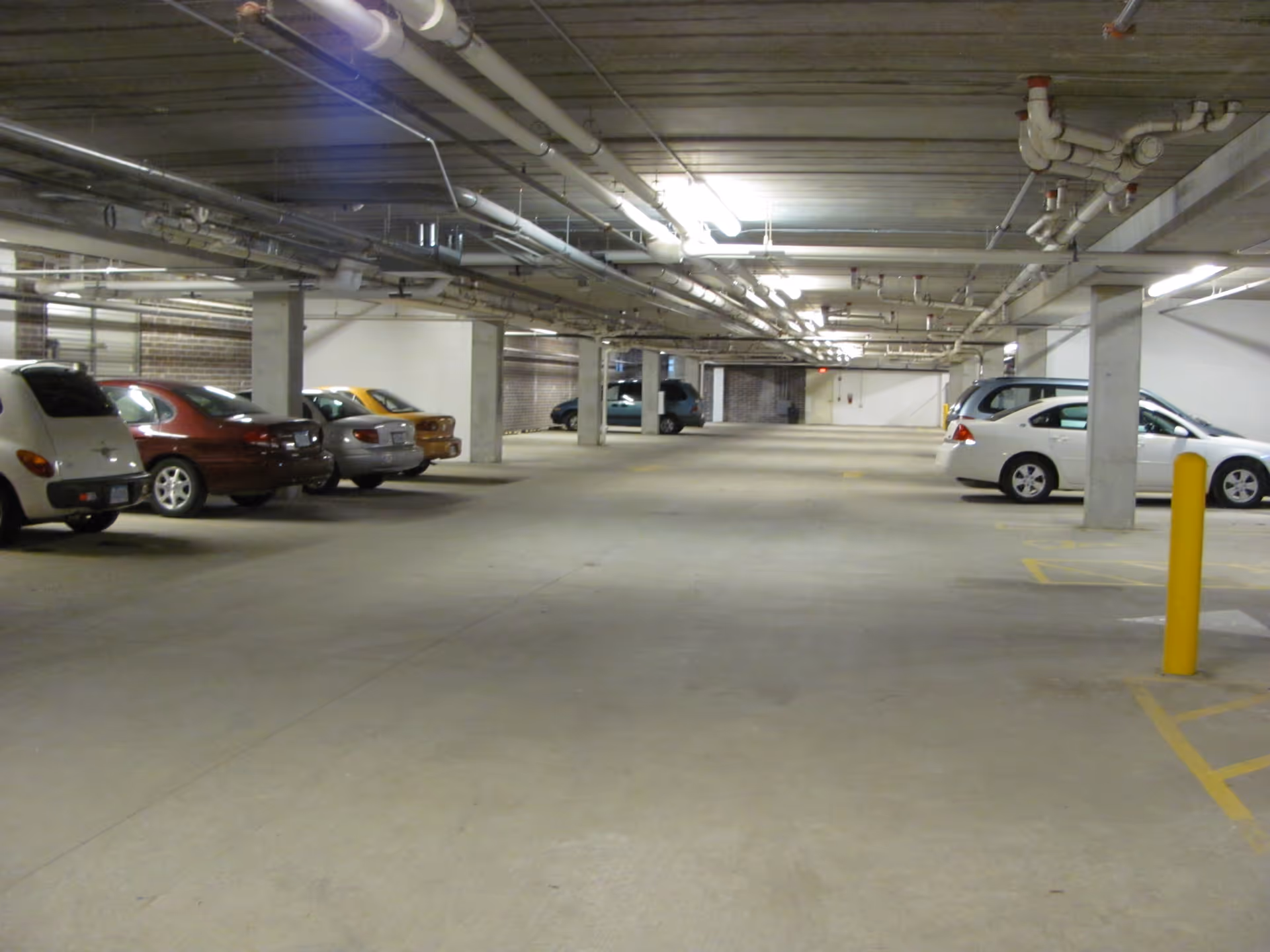 Indoor parking garage with several parked cars along the sides and concrete pillars supporting the ceiling with exposed pipes and lighting fixtures.