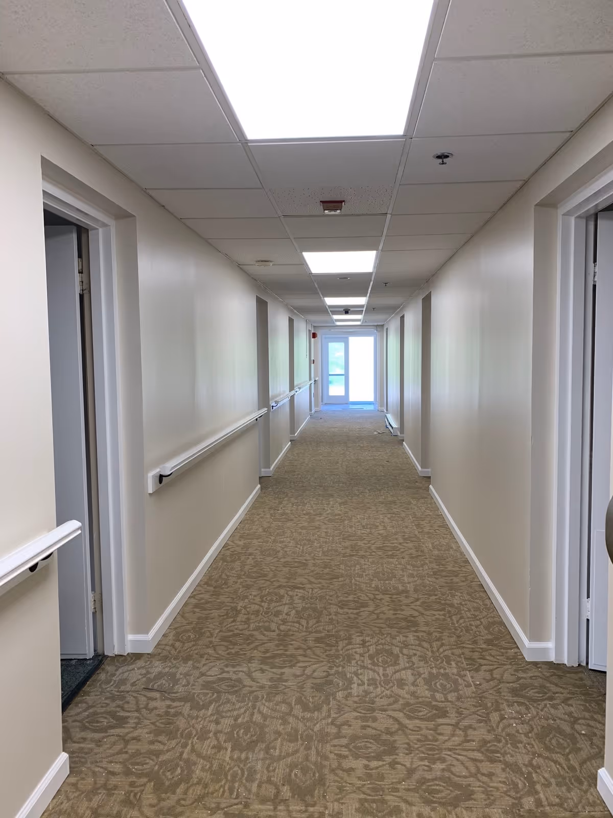 A long, empty hallway in a senior living facility with beige patterned carpet, cream-colored walls, white handrails on both sides, and several open doorways leading to rooms. The ceiling has recessed fluorescent lighting panels, and a glass door is visible at the far end of the corridor.