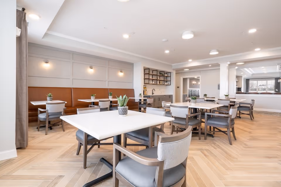 Spacious bright dining room with white tables, gray chairs, booth seating along the wall, and small potted plants.