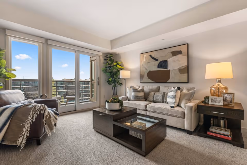 A bright living room with a beige sofa adorned with multiple cushions, a dark wooden coffee table with decorative items, a side table with a lamp and framed photos, a leather armchair with a throw blanket, large windows and a glass door leading to a balcony with outdoor seating, and a large abstract painting on the wall.