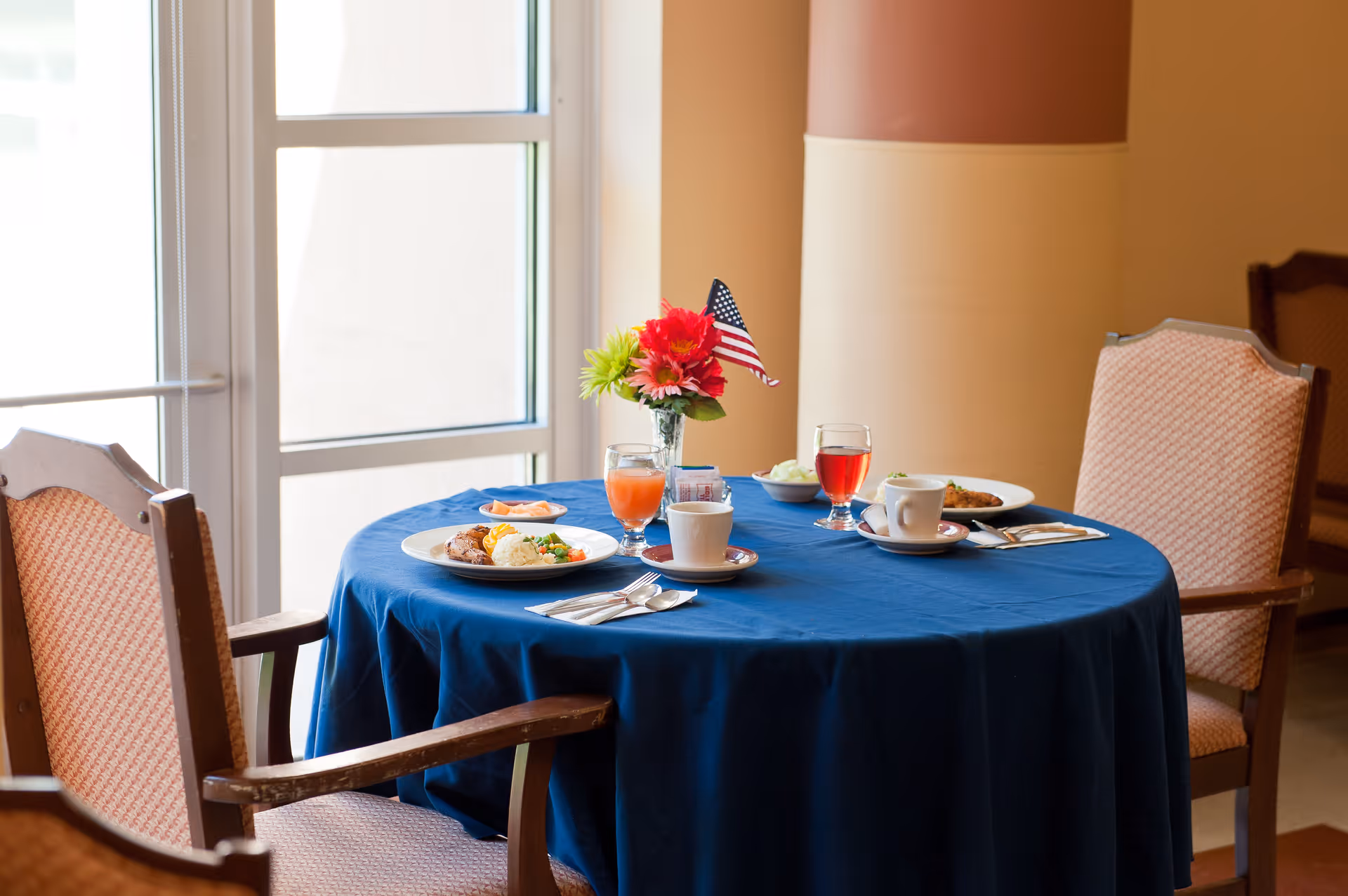 Round dining table with a blue tablecloth set with plates, drinks, cups and a small vase of flowers with an American flag, surrounded by upholstered chairs near a window.