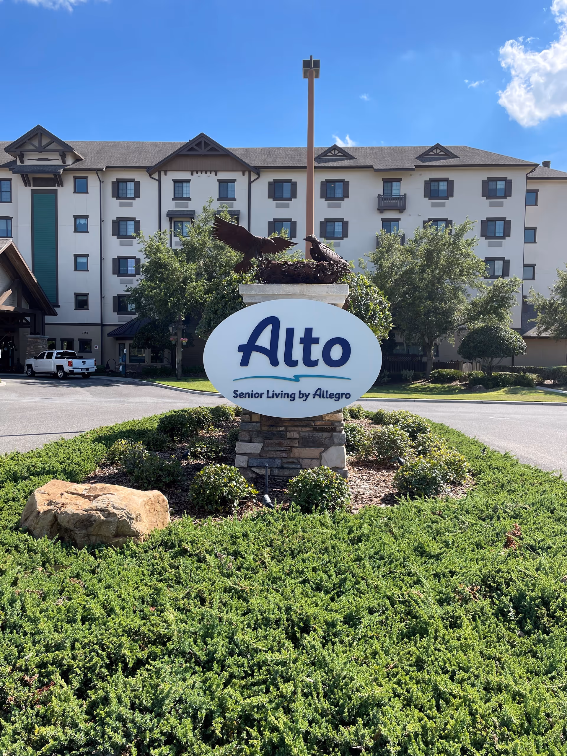Exterior view of Alto Senior Living by Allegro facility in Tavares, FL, showing a multi-story building with multiple windows and a landscaped roundabout with a stone pillar and a sign that reads 'Alto Senior Living by Allegro' topped with a sculpture of two birds.