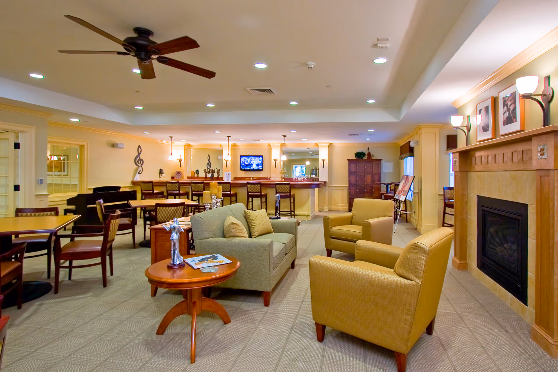 Spacious senior living common room with sofas and armchairs around a coffee table, dining tables and a bar in the background and a fireplace on the right.
