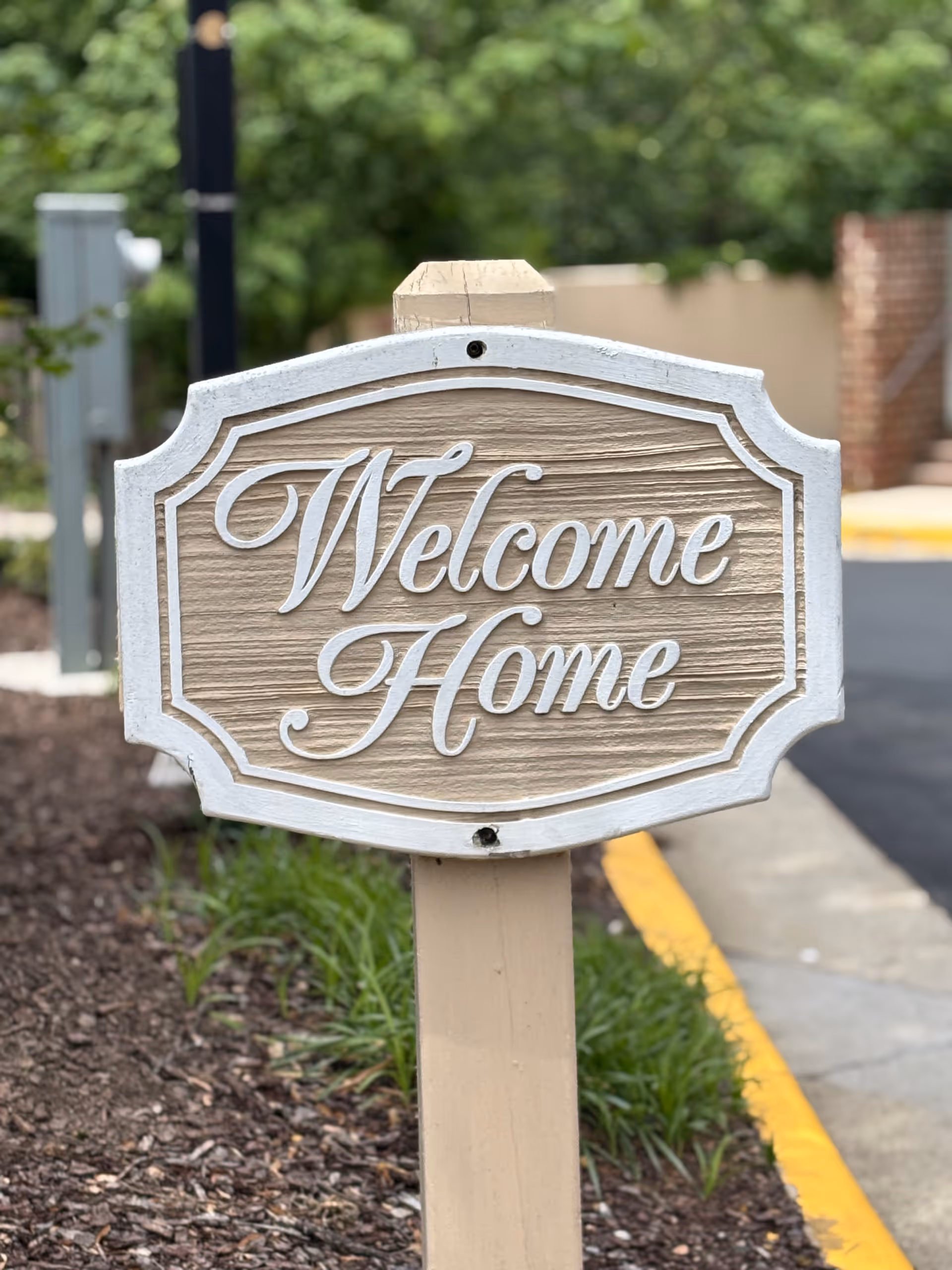 A wooden sign with white trim and cursive white lettering that reads 'Welcome Home' mounted on a beige post, surrounded by mulch and greenery with a blurred background of a sidewalk and street curb.