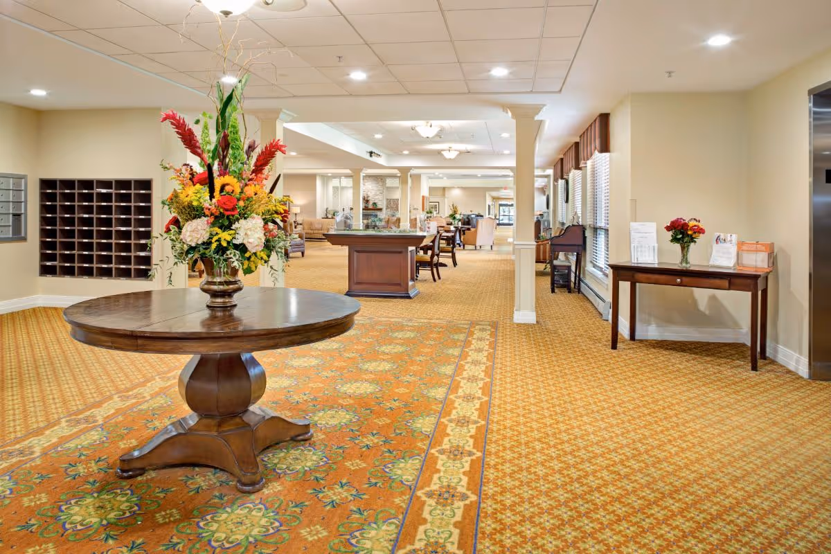 A spacious and well-lit interior common area of a senior living facility with patterned carpet, a round wooden table with a large floral arrangement in the center, mailboxes on the left wall, and seating areas with chairs and tables in the background. There is also a small table with flowers and brochures near a window on the right side.