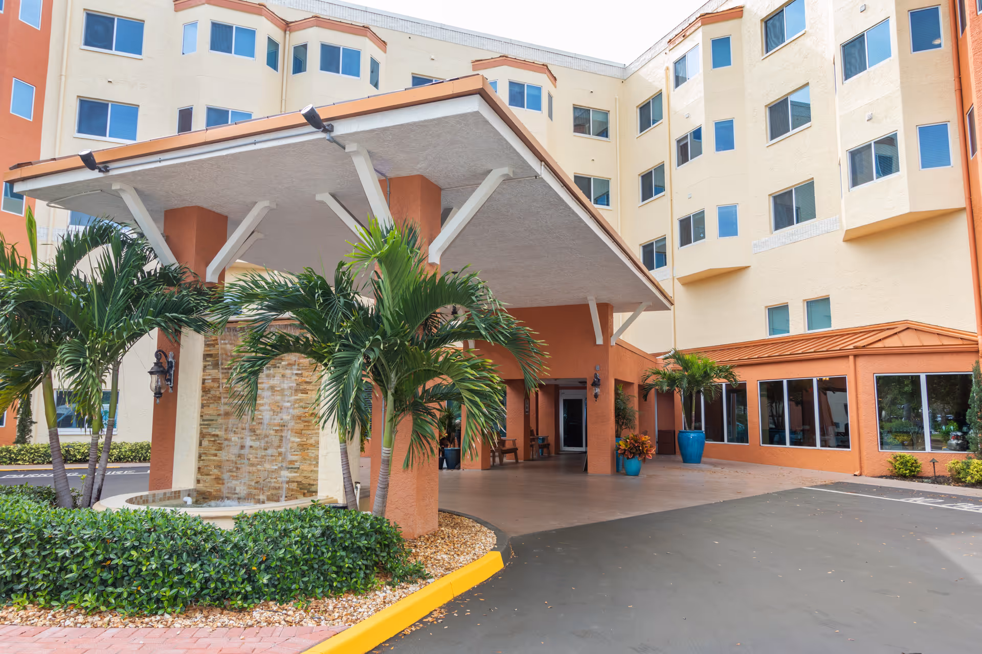 Entrance area of a multi-story senior living facility with a covered drop-off zone, palm trees, and landscaped greenery. The building has beige and orange exterior walls with multiple windows.
