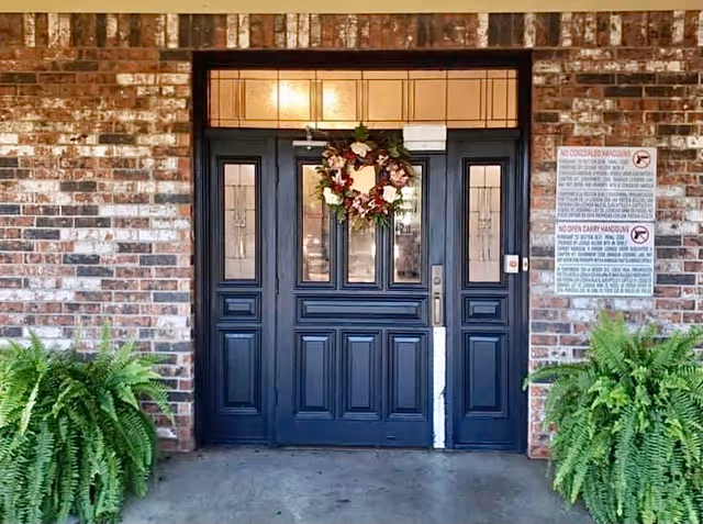 Dark paneled double entrance doors with a floral wreath set in a brick facade flanked by potted ferns.